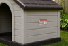 A plastic dog house with a light beige siding and a dark gray shingled roof, situated on a green lawn. A red and white warning sign on the side reads 'DOGS' with a red cross symbol.