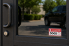 A black metal security door with a glass window showing a blurred outdoor scene and a black SUV reflection. A white and red emergency sign on the bottom right reads 'IN CASE OF EMERGENCY PLEASE RESCUE MY: DOGS'.
