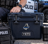 A black YETI hard cooler with a thin blue line police dog sticker on the front, shown outdoors next to a police officer's hand, a tactical backpack, and a black YETI tumbler.